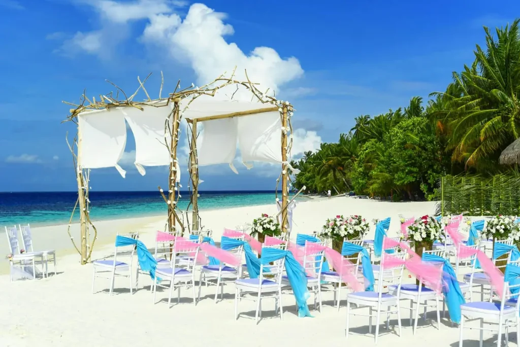 Ceremonia de boda en la playa con arco de madera y tela blanca, sillas blancas decoradas con lazos azul turquesa y rosa, y palmeras tropicales de fondo en el Caribe