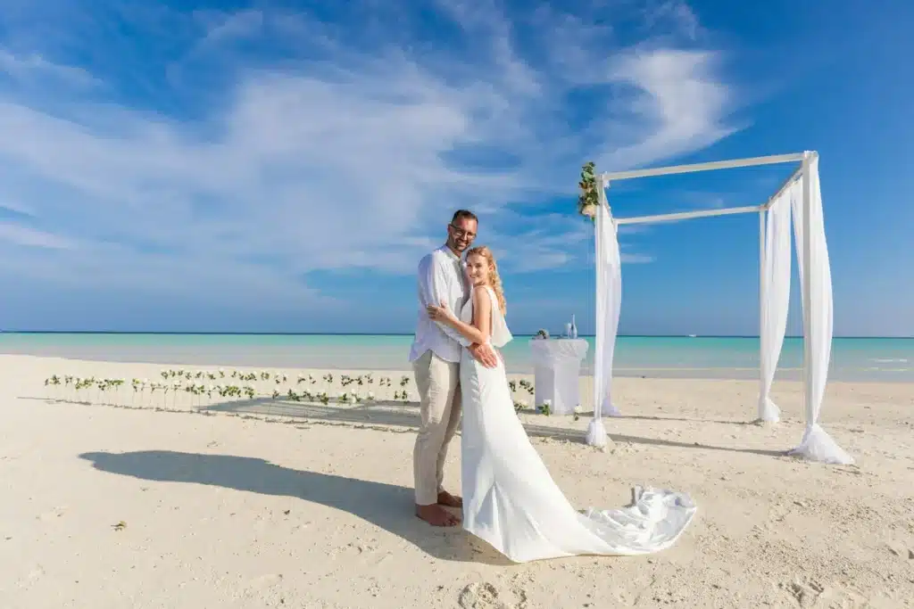 Pareja de recién casados abrazada frente a un arco blanco en la playa de arena blanca con el mar turquesa del Caribe de fondo en Playa Mujeres